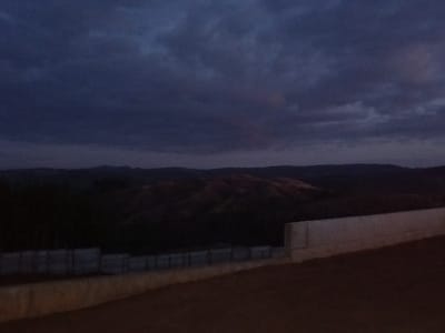 The image depicts a rural or semi-rural landscape in Santa Juana, Chile, at dusk or during nighttime under overcast conditions. The sky, occupying the upper two-thirds of the frame, is a dark blue-grey with layered cloud formations. A distant horizon line reveals multiple dark, undulating hills or mountain ranges. In the mid-ground, patches of lighter, reddish-brown earth are visible on some hill slopes, suggesting exposed soil. Below the distant hills, a dark, vegetated area or terrain is present. In the lower left quadrant of the image, a horizontal series of light grey, possibly concrete, fence panels or sections of a low retaining wall are visible. The foreground is composed of a prominent light grey concrete retaining wall that angles from the lower right corner upwards and to the left, terminating near the center-left. Adjacent to and in front of this wall, the ground consists of a dark reddish-brown earthen or dirt surface that slopes slightly downwards from right to left. No individuals, animals, or explicit actions are present in the scene.