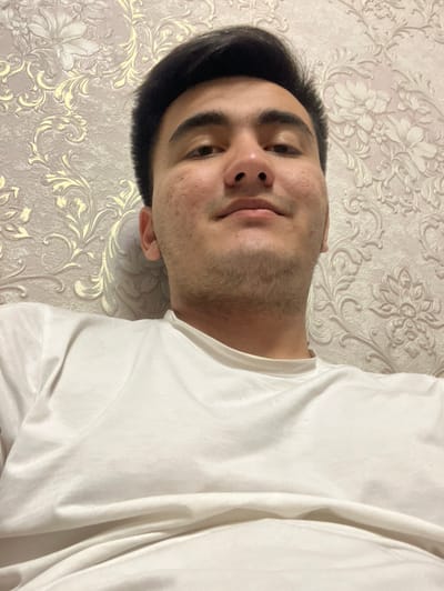 A close-up, upward-angled photograph depicts a young adult male, likely self-captured, in an interior setting in Namangan City, Uzbekistan. The subject has dark hair, a light complexion with visible facial stubble and minor skin texture, and wears a plain white crew-neck t-shirt. He faces the camera directly with a neutral expression. The background wall is covered in light-colored wallpaper displaying a repetitive, ornate floral and scroll pattern in a muted golden or beige hue. No external identifying features are present to determine a specific location within the city beyond an indoor environment.