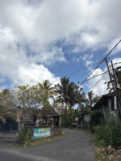 A residential street in Tampaksiring, Indonesia, is depicted under a partly cloudy sky. The road curves to the right, lined with vegetation, palm trees, and low-rise buildings. On the left, a building with a weathered facade and a tiled roof is adorned with a colorful banner that reads "V.S.L PLAYGROUP." A sign with text is visible on a pole in front of this building. To the right, another building with a similar rustic aesthetic features a sign with Indonesian text. Utility poles with numerous wires are prominent in the background, stretching across the scene. The overall impression is that of a tropical, rural setting within a town.