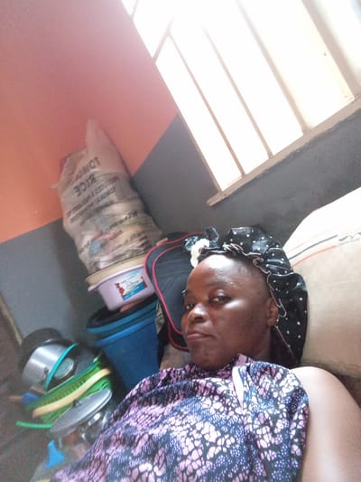 The image depicts an indoor scene, likely a residential room in Kason, Nigeria. A woman is lying down, partially visible. She is wearing a black bonnet with white polka dots and a pink and blue patterned top. Her facial features are distinct, and she appears to be looking towards the camera. In the background, there are various objects including stacked pots, a blue bucket, and a large bag of rice. The lighting suggests natural light filtering through a window with blinds. The overall environment appears to be a cluttered living space.
