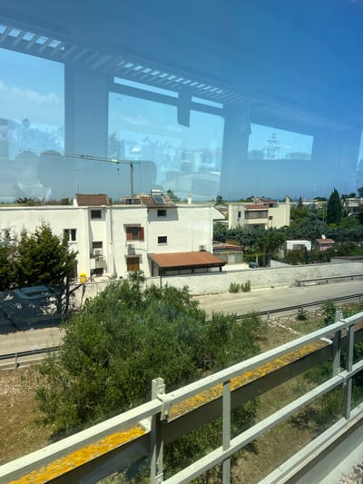 This image shows a view from inside a vehicle, looking out through a window at a suburban area in Bari, Italy. The scene is dominated by buildings and vegetation, with a notable reflection of an overhead structure, possibly a bridge or overpass, on the window.
In the foreground, a metal railing with yellow markings runs horizontally across the bottom of the frame. Beyond the railing is a grassy area and a road. Further back, a low white wall with a textured surface separates the road from more vegetation and buildings.
The central focus of the image is a cluster of white buildings with terracotta roofs. One of these buildings is a two-story structure with several windows, air conditioning units, and solar panels on the roof. Adjacent to it are other houses, some with balconies and awnings, surrounded by lush green trees and bushes. A construction crane is visible in the background, suggesting ongoing development in the area.
The reflection on the window adds a layer of complexity to the image, showing the underside of an architectural structure with parallel beams. The sky is blue with scattered clouds. The overall impression is one of a sunny day in a residential area with a mix of modern and older construction. The specific location within Bari is not identifiable from the visual cues alone.