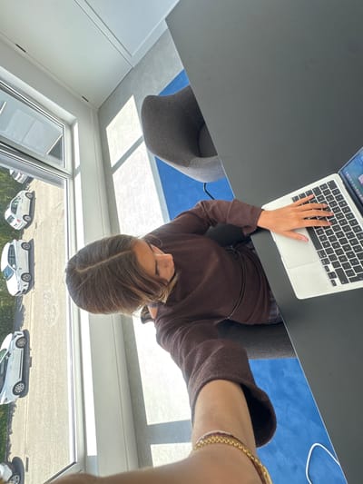 The image, rotated 90 degrees counter-clockwise, captures a young person focused on a laptop in a bright, modern indoor setting, likely an office or showroom, in Torres Vedras, Portugal.
The subject, with light brown hair, is seen from an overhead perspective, wearing a dark brown long-sleeved top. They are seated at a dark, minimalist desk, their right hand actively typing on the QWERTY keyboard of a silver laptop. The laptop screen displays a desktop interface with various application icons. A grey upholstered chair is partially visible under the desk. A gold chain bracelet and a multi-strand bracelet adorn the person's right wrist, which is visible in the foreground, suggesting a selfie from a low angle.
The immediate surroundings include the dark desk surface and, underneath, a section of blue flooring or carpet with a white power cord. The background features a large window or glass wall, through which several white cars are parked outdoors, possibly in a display area or parking lot, bordered by green foliage. The interior ceiling has multiple rectangular light panels, indicating ample natural light during the daytime. The presence of the 'Ç' key on the laptop keyboard aligns with the given location context of Portugal. The scene conveys a sense of focused work or study in a clean, contemporary environment.