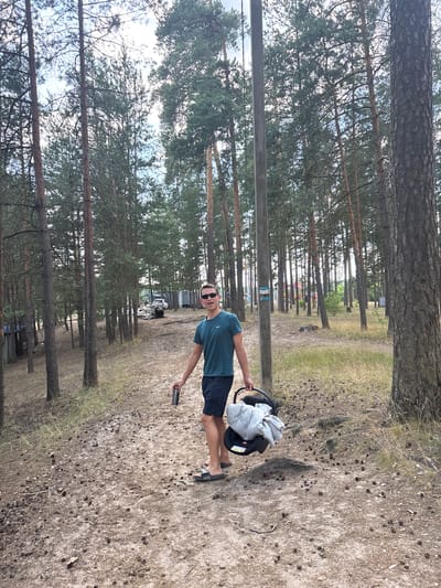 A young man with sunglasses and a green t-shirt is standing on a sandy path in a pine forest. He is holding a can in his right hand and a baby carrier with a gray blanket in his left. The path is littered with pine cones. In the background, there are several pine trees, a wooden pole with a sign, and some buildings and vehicles. The sky is overcast. The location is the city of Kurovskoye, Russia.