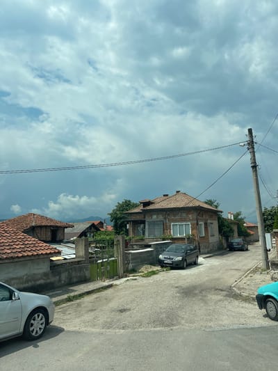 This image captures a static street scene in the town of Karnare, Bulgaria, under an overcast sky filled with dense, light grey clouds.
The street itself is a narrow, unpaved or poorly maintained asphalt road, exhibiting a mix of dirt, gravel, and numerous potholes and uneven surfaces, indicating disrepair. It appears to curve slightly to the right and may have a gentle incline.
On the left side of the foreground, the front-left quarter of a silver Volkswagen Golf is visible. Further down the street on the left, a dark grey Toyota Corolla with Bulgarian license plate B A8761BH is parked beside the road. Adjacent to the road are various residential structures. In the mid-ground left, a house with a reddish-brown terracotta tiled roof is partially seen, and behind it, other buildings with varying roof types, including corrugated metal and older tiles, are present. A weathered green metal gate, flanked by concrete posts, leads into a property.
On the right side of the image, a tall concrete utility pole with multiple cross-arms supporting electrical wires is a dominant feature. A single street light fixture is mounted on the pole. Multiple black wires extend from the pole across the street, creating linear elements in the upper mid-ground. Behind the utility pole and further along the street, a prominent two-story house with a reddish-brown tiled roof and a brick facade, showing signs of wear and aging, features several windows including a large multi-paned window on the lower level. A dark sedan, possibly a Mercedes-Benz, with a partially visible Bulgarian license plate B 4251KM, is parked in front of this house, facing away from the viewer.
In the background, beneath the heavy cloud cover, distant hills or mountains are faintly discernible. Scattered trees and shrubs are visible around the houses, providing limited greenery to the otherwise largely concrete and brick environment. No people are present, and no actions are depicted.