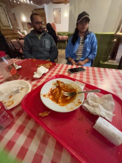 The image shows a table inside a restaurant, likely an Italian restaurant, in Bradford, UK. The table is covered with a red and white checkered tablecloth. Two people are seated at the table. A man wearing a blue jacket and a woman wearing a blue shirt and a baseball cap. There are two empty plates, one with remnants of pasta, and a red tray containing a partially eaten plate of pasta. A fork is on the empty plate. A can of soda and a receipt are also on the table. Several other tables and chairs are visible in the background, with other diners. The atmosphere appears to be casual and relaxed. The restaurant has a simple and rustic interior. There are no visible windows in this room, making it difficult to determine the exact location within the city.