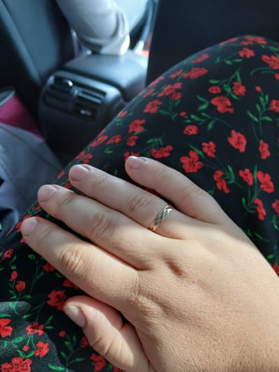 A close-up shot focuses on a woman's hand resting on a floral-patterned fabric. The fabric is black with red floral designs and green stems. The woman is wearing a thin, textured silver ring on her left ring finger. Her hand is positioned to the left of the frame, with fingers slightly curled. The background is blurred, showing the interior of a vehicle. A dark car seat and a rear air vent are visible, suggesting the scene takes place within a car. The lighting indicates it is daytime, with sunlight casting shadows on the hand. The image does not depict anything that would indicate the specific location of Sorochkino, Russia.