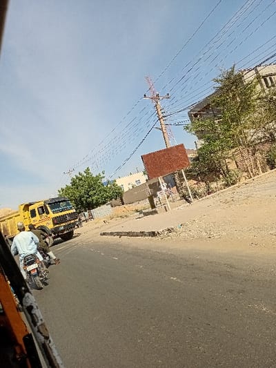 The image portrays a daylight street scene in Sharada, Nigeria, captured from a low, tilted perspective, with a partial view of an orange/yellow vehicle frame in the lower-left foreground. An asphalt road, featuring a faint white dashed line, occupies the central midground. A person wearing a light blue garment and a cap operates a black and white motorcycle, traveling away from the viewer, with a second individual in dark clothing riding as a passenger. To the left, a yellow dump truck with a black cab is visible. On the right, a concrete curb delineates the road from a dirt and gravel shoulder populated by green vegetation. In the background, a multi-story, light-colored building with dark windows stands on the right. A tall, reddish-brown utility pole, supporting numerous overhead wires, is prominently positioned on the right, adjacent to a large, rectangular, reddish-brown sign board mounted on posts, facing away from the camera. The sky is clear blue.
