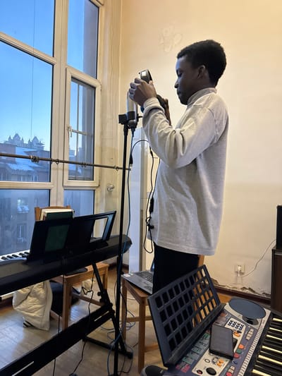 An image taken indoors, likely a music production space, features a young dark-skinned male in profile, facing right, adjusting black headphones with both hands. He is wearing a light grey long-sleeved turtleneck shirt and dark pants. To his immediate left is a silver condenser microphone on a black stand. The room has light-colored walls with visible peeling paint and a large multi-paned window on the left side, showing an urban landscape. Outside, under a clear blue sky, buildings with light-colored or snow-dusted rooftops are visible, and in the distance, a distinctive, ornate building with multiple turrets or peaked roofs stands out, characteristic of certain architecture found in Shenyang City, China.

In the foreground to the left, a black electronic keyboard with a music stand holding several books is positioned. A white puffy jacket is draped below it. Further right, closer to the viewer, is another, older model electronic keyboard, light-colored, with a black grid-like accessory and a dark rectangular object on its surface. A silver and black laptop rests open on a wooden chair to the right of the first keyboard, positioned beneath the male subject. Various audio cables run along the floor and up the equipment stands. An electrical outlet is visible on the right wall. The scene captures a moment of preparation or active engagement with audio equipment within a modestly appointed room in Shenyang City, China.