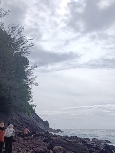 The photograph depicts a rocky shoreline in Naga Umbang, Indonesia, under a cloudy sky. The foreground features large, dark rocks along the beach. In the middle ground, several people are present. Two individuals, one wearing an orange shirt and the other a headscarf, stand near the left edge of the frame. A third person sits on the rocks further to the right. In the background, ocean waves crash against the rocky shore. A vegetated cliff rises sharply on the left side of the frame.