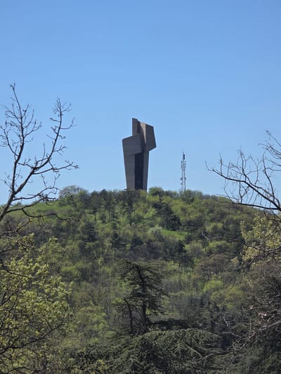 The image depicts a concrete monument in Pobeda, Bulgaria, against a blue sky. The monument has a geometric shape and is positioned near a hilltop covered with green trees. In the background, a transmission tower is visible. Leafless tree branches are prominent in the foreground, framing the monument and the landscape. The image is oriented vertically.