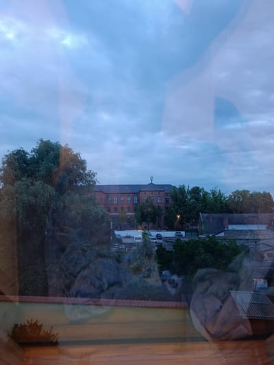 The image depicts a view through a window, showing a reflection superimposed on the exterior scene. The exterior scene features a large, brick building with arched windows, likely a former industrial or institutional building, situated in Roßlau, Germany. To the left of the building, a dense cluster of trees with dark green foliage is prominent. Beyond the main brick structure, other buildings, including a house with a gabled roof and a smaller building with a corrugated roof, are visible. Several cars are parked in front of the brick building, and a caravan is also present. The sky is overcast with grey clouds. The reflection on the window includes abstract shapes and colors, obscuring parts of the exterior view. The lower part of the image shows a wooden surface and a reddish tiled structure, possibly a windowsill and a roofline.