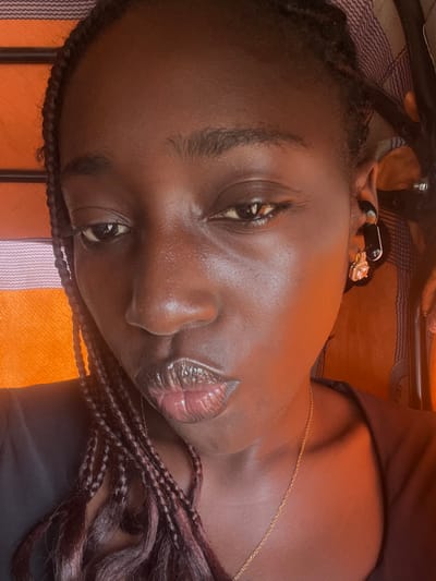 A close-up, head-and-shoulders portrait of a young Black woman with braided hair and dark skin. She is wearing a black top, a delicate gold chain, and earrings. One earring is a black stud, and the other is a gold hoop with a clear, faceted stone. Her lips are slightly parted, and her gaze is directed towards the viewer. The background features an orange and striped fabric. The lighting appears to be natural, casting subtle shadows and highlighting the sheen on her skin. The overall impression is one of a casual, personal photograph. The location within Karum, Nigeria, is not specified in the image itself.