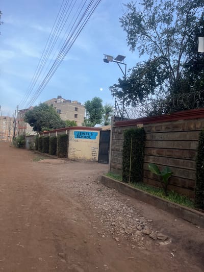 This image, captured in Gitothua ward, Kenya, presents a static outdoor scene, likely during early morning or late afternoon, characterized by a bright, clear blue sky and a visible moon. The foreground is dominated by a dirt road, uneven and scattered with gravel and small rocks, particularly along its right side. A concrete curb separates the road from a neatly trimmed green hedge, positioned in front of a stone-patterned wall. This wall, topped with what appears to be barbed or razor wire, suggests security.
An entrance in the wall features a dark-colored metal gate. To its left, a blue and yellow sign is attached, prominently displaying "JEWELS SCHOOL." Below this, "Mother Foundation" is partly visible, along with academic levels: "PP1," "PP2," and "Grade 1."
Overhead, a streetlight with visible solar panels extends from a pole. Multiple power lines or utility cables stretch diagonally across the upper left sky. Lush green trees are visible, some overhanging the scene, with more foliage and several multi-story buildings in the background, hinting at a residential or institutional area. The architecture of the buildings is functional, contributing to an urban-fringe atmosphere. No explicit activity or people are present, offering a tranquil view of the local environment.