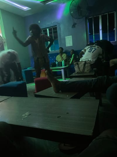 A dimly lit room with a group of people. Several people are seated at tables, some facing computer screens. A figure stands in the center, appearing to dance or move energetically. The room has a dark blue hue with green and purple lighting. There's a fan on the wall, windows, and a bag on the table near a person wearing a sports jersey with the number 97.