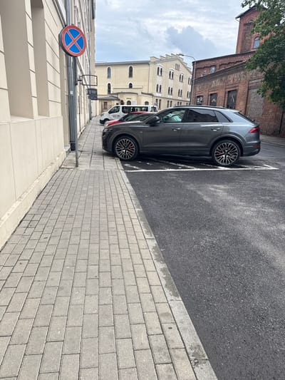 The image shows a street scene in Zielona Góra, Poland. The perspective is from a low angle, looking up towards a gray SUV parked on the right side of the frame, which is oriented vertically. The car is parked in a designated spot marked by white lines on asphalt. In the background, there are several buildings: a red brick industrial building on the left, a cream-colored building with arched windows in the middle ground, and a white van is visible behind the SUV. A street sign with a red circle and a blue cross inside is visible on the left, indicating a prohibition. The weather appears to be overcast, with a cloudy sky. The ground is paved with light gray bricks, and there is a concrete sidewalk. The overall scene depicts an urban environment, likely during the daytime. No people are visible in the image. The visible text on the white van is "EURO-WIEL". There is also text on a sign in the background which is difficult to read clearly.
