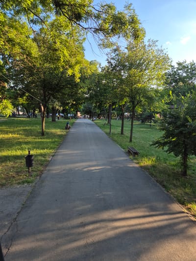 A paved walkway extends through a park with lush green grass and numerous trees. The sun casts long shadows across the path, indicating it is late afternoon or early evening. Several individuals are visible in the distance, walking or sitting on benches. A man in a blue shirt is seated on a bench on the right side of the path, facing away from the camera. Another man is visible in the distance walking along the path. There are also playground structures visible in the background to the left. The overall scene depicts a serene park environment in the city of Gradska opština Crveni Krst, Serbia.