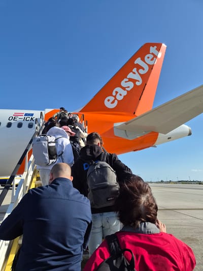 Passengers are boarding an EasyJet airplane, evidenced by the tail fin displaying the company logo. The aircraft is marked with "OE-ICK," indicating its registration. Passengers are ascending the stairs towards the plane's entrance. The location may be the city of Moreira, Portugal, but this is impossible to ascertain from the image alone. The day is clear with a blue sky. The apron is visible.