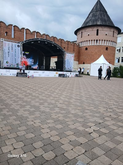 This media file depicts an outdoor event in Tula, Russia, with a large brick tower in the background, likely a historical fortress. In the foreground, a stage is set up with a large screen displaying the text "Молодёжи 2026" (Youth 2026) and "ПОДЁЖИ" (likely part of a word related to youth or the event name). The stage area has banners and branding for various sponsors, including "HAIRURG," "MAMA MIA," and "К-ХОЛДИНГ."
Several people are present. Two individuals in black clothing are standing near a white tent, seemingly involved in organizing or supervising the event. A group of people in white shirts and dark pants are also visible, possibly participants or staff. Closer to the stage, a woman and a child are interacting with a scooter. The overall atmosphere appears to be a public gathering or festival, possibly during daytime given the lighting. The ground is paved with patterned bricks. The sky is overcast, suggesting a cloudy day. The scene captures a moment of a community event, likely a festival or a youth-focused gathering, with strong branding and a historical backdrop.
