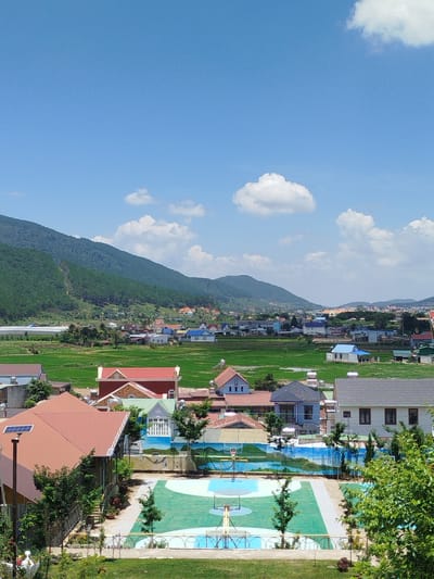 This is a high-angle, full-shot photograph taken in the city of K'Rèn, Vietnam, under a bright, blue sky with scattered white clouds. The image depicts a rural or semi-rural residential area nestled against a backdrop of lush green mountains. In the foreground, a multi-colored basketball court is visible, with a green playing surface and blue and white markings. Several trees are planted around the court. Beyond the court, a collection of houses with red and gray roofs are situated. Further in the distance, more houses are scattered across the landscape, interspersed with vibrant green fields, likely rice paddies. The mountains in the background are covered in dense green foliage. A greenhouse structure is visible on the left side of the frame in the mid-ground. There are no people or animals clearly visible in the image. The overall scene conveys a sense of peaceful, rural life with a clear, sunny day.