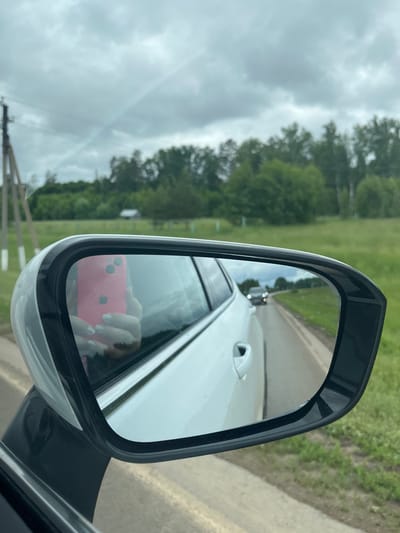 The image depicts a view from the interior of a white vehicle, primarily focusing on its right-hand side exterior mirror and the surrounding landscape. Reflected in the mirror is a human hand with light-colored nail polish, holding a pink smartphone with an identifiable Apple logo and dual camera lenses, indicating the photographer. The reflection also shows the white body of the car. Through the mirror, a two-lane asphalt road extends into the distance, with a dark-colored vehicle approaching, its headlights illuminated. The surface of the mirror is dotted with water droplets. Directly visible outside the vehicle, to the left of the mirror, is the white door panel and handle of the car. The immediate roadside features a grassy area. In the midground, a line of green deciduous trees forms a natural barrier, with a small, light-colored structure partially visible beneath them. To the far left, a wooden utility pole is partially present. The sky above is entirely overcast with grey clouds, displaying faint, intersecting linear patterns consistent with aircraft contrails. The scene is located in Бизяки, Russia.