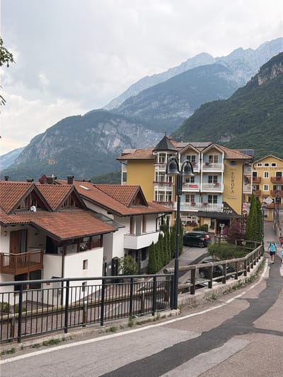 In the mountainous region of Molveno, Italy, a street scene unfolds. The foreground features an asphalt road with white markings and a shadow cast across it. A metal railing borders the road, beyond which lies a retaining wall constructed of concrete and wood. Several parked cars are visible, including a black SUV and a darker vehicle. To the left, residential buildings with red tile roofs and white facades are nestled against the mountainside. Further to the right, a multi-story yellow hotel named "HOTEL EUROPA" stands prominently, adorned with balconies and flower boxes. The background is dominated by a majestic mountain range, its slopes covered in dense green foliage, partially obscured by a cloudy sky. Two individuals are walking away from the camera on the right side of the road, one wearing a blue shirt and shorts, and the other a light-colored top and shorts. A person is also visible near the entrance of the hotel.