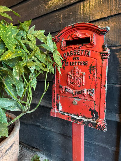 A close-up, eye-level shot shows a weathered, bright red mailbox with peeling paint, mounted on a red post, standing against a dark, horizontally-planked wooden wall in Eyüpsultan, Turkey. The mailbox features ornate detailing, including the embossed words "CASSETTA PER LE LETTERE" and a crest with the words "REGIE POSTE" below it. A small keyhole is visible near the bottom. To the left of the mailbox, a potted plant with green leaves speckled with yellow is visible, partially obscuring the mailbox. The overall impression is one of age and character, suggesting a historical or vintage setting.