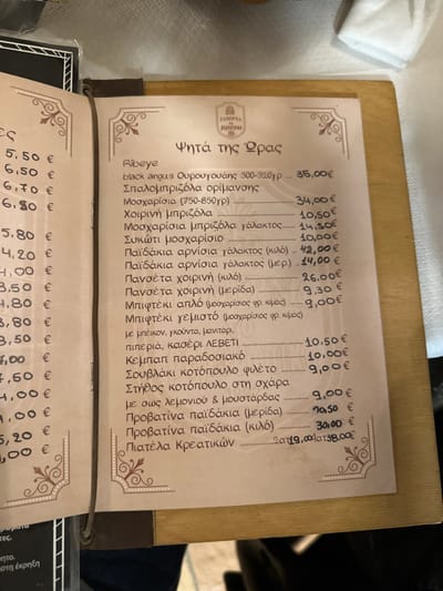 The image displays an open menu from a Greek restaurant, presented on a wooden surface, likely a table or a menu holder. Part of a white tablecloth is visible in the upper left corner, and a corner of another menu is seen at the bottom left.

The activity taking place is the presentation of the restaurant's grilled dishes menu. The image is taken at an angle, making the text appear rotated clockwise.

Notable details include the ornate decorative borders on the menu page, lending a traditional or classic aesthetic. The prices are listed in Euros. The lighting suggests an indoor restaurant setting.

Visible text on the main menu includes the title "Ψητά της Ώρας" (Grilled Dishes of the Hour/Season) and a logo that reads "ΤΑΒΕΡΝΑ ΠΕΡΙΟΥΣΙΚΟ" (Taverna Periousiko). The menu lists various meat dishes and their prices:
- Ribeye black angus Ουρουγουάης 300-320γρ. ... 35,00€
- Σπαλομπριζόλα ορίμανσης Μοσχαρίσια (750-850γρ) ... 34,00€
- Χοιρινή μπριζόλα ... 10,50€
- Μοσχαρίσια μπριζόλα γάλακτος ... 14,50€
- Συκώτι μοσχαρίσιο ... 10,00€
- Παϊδάκια αρνίσια γάλακτος (κιλό) ... 42,00€
- Παϊδάκια αρνίσια γάλακτος (μερ.) ... 14,00€
- Πανσέτα χοιρινή (κιλό) ... 26,00€
- Πανσέτα χοιρινή (μερίδα) ... 9,30€
- Μπιφτέκι απλό (μοσχαρίσιος φρ. κιμάς) ... 9,00€
- Μπιφτέκι γεμιστό (μοσχαρίσιος φρ. κιμάς) με μπέικον, γκούντα, μανιτάρι, πιπεριά, κασέρι ΛΕΒΕΤΙ ... 10,50€
- Κεμπαπ παραδοσιακό ... 10,00€
- Σουβλάκι κοτόπουλο φιλέτο ... 9,00€
- Στήθος κοτόπουλο στη σχάρα με σως λεμονιού & μουστάρδας ... 9,00€
- Προβατίνα παϊδάκια (μερίδα) ... 10,50€
- Προβατίνα παϊδάκια (κιλό) ... 30,00€
- Πιατέλα Κρεατικών (Meat Platter) with a handwritten "2 ατομα" (2 people) and a price of 38,00€.
The partially visible menu on the left also shows prices, ranging from 1,50€ to 9,00€.
