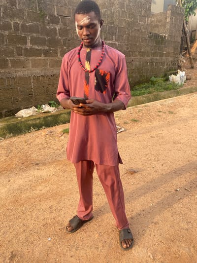 A man wearing a maroon-colored, long-sleeved tunic and matching trousers is standing on a dirt road in Afikpo, Nigeria. He is wearing black sandals and a beaded necklace. He is holding a black smartphone in his hands. To his left is a gray brick wall. Between the wall and the dirt road is an area of grass and weeds. There is trash on the ground near the wall. The dirt road extends into the background. The light is bright, suggesting it is daytime.