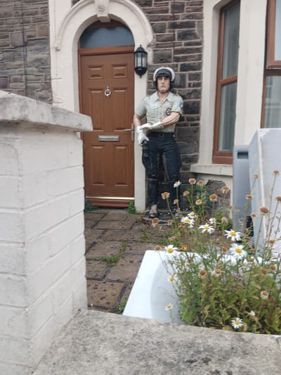The image shows a garden gnome dressed as a police officer standing outside a terraced house in Bristol, United Kingdom. The gnome is wearing a white helmet, a light-colored shirt with a badge, black trousers, and black boots. It is standing to the left of a brown wooden front door with a silver letterbox and a silver door knocker. To the right of the door, there is a window with a brown frame. In front of the gnome and the house, there are several daisy flowers in a white bathtub, which appears to be used as a planter. The house has a brick facade. The foreground is occupied by a concrete wall. The scene is outdoors, likely on a slightly overcast day.