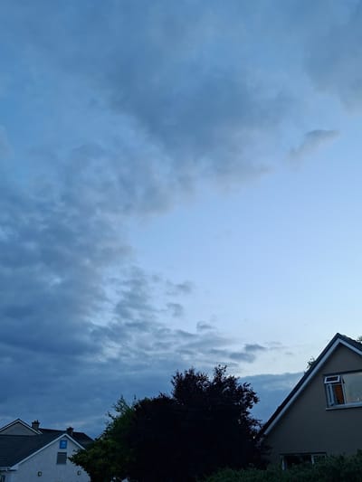 The image depicts a cloudy sky over a residential area in the city of Galway, Ireland. In the foreground, the roofs of several houses are visible. One house on the left has a grey tiled roof with a white facade and a blue sign with "JW ORG" written on it. To the right, another house with a dark grey roof and a light brown facade features a visible window with an open pane reflecting a warm light. Dense foliage, including dark-leaved trees, obscures parts of the lower half of the image. The sky is predominantly filled with grey and blue clouds, with a lighter blue sky visible towards the upper right. The lighting suggests it is either early morning or late evening.