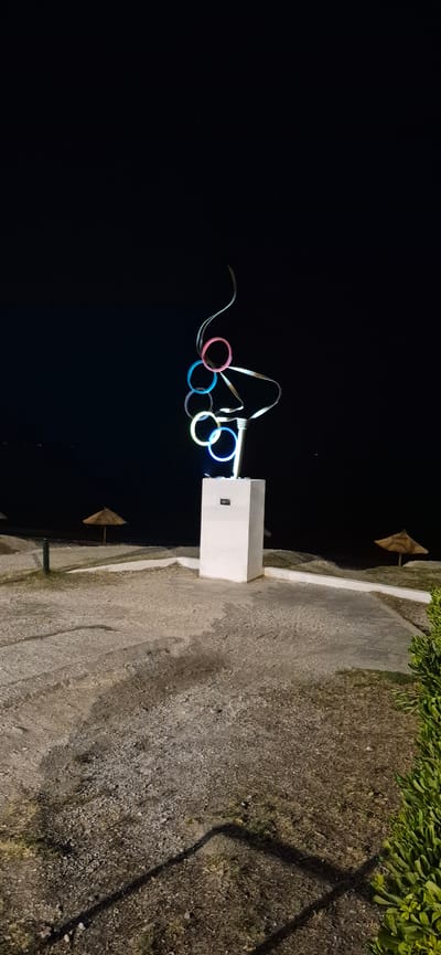 A night-time, eye-level, vertical shot captures a sculpture of Olympic rings and wavy metallic strands in Paleo Faliro, Greece. The sculpture is illuminated by neon lights in pink, blue, and yellow, and is mounted on a white rectangular pedestal. The background is a dark night sky, with a faint glimmer of distant lights on the horizon. To the left and right of the sculpture, two thatched beach umbrellas are visible on a gravelly and paved area. A dark green shrub is situated on the right edge of the frame. The scene is devoid of any people or animals.