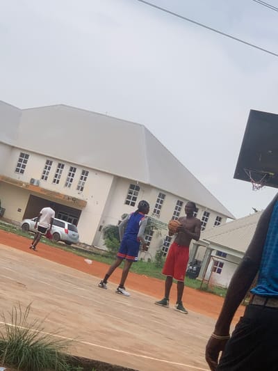 In Ukukwa, Nigeria, two young men are engaged in a basketball game on an outdoor court. One player, shirtless and wearing red shorts, holds a basketball, preparing to shoot or pass. He is positioned to the right of the frame. To his left, another player, dressed in a blue and orange basketball uniform, appears to be on defense, anticipating the next move. The court is made of a reddish-brown material, with white lines marking its boundaries.
Behind the court, a large, cream-colored building with a white, angled roof dominates the background. The building has numerous windows, suggesting it might be a school or administrative facility. A silver car is parked near the building, partially visible. The sky is overcast, indicating a cloudy day, and the lighting suggests it is daytime. The atmosphere seems casual and energetic, typical of a friendly game.
The image is rotated, with the subjects appearing on a diagonal. The overall scene depicts a community gathering or recreational activity taking place in an outdoor urban or semi-urban environment in Nigeria.
