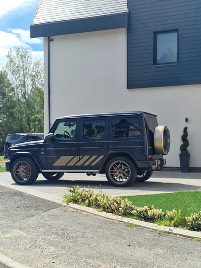 A black Mercedes-Benz G-Wagon with gold accents is parked on a paved driveway in front of a modern house. The house has a light-colored exterior wall and a dark blue vertical paneling section with a square window. There's a patch of green lawn between the driveway and the house, and a neatly trimmed evergreen topiary in a black pot stands next to the house. The location is Witton Gilbert, United Kingdom. The lighting suggests it's daytime.