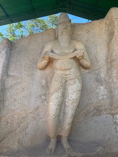 The image shows the Gal Vihara reclining Buddha statue, located in Polonnaruwa, Sri Lanka. The statue is carved into a large rock face. The Buddha is depicted lying on his right side, with his head resting on his right hand. His left hand is extended along his body. The sculpture exhibits fine details and a serene expression. The surrounding rock surface is textured. A green corrugated roof structure is partially visible on the left edge of the image, possibly indicating a protective shelter. Trees and the blue sky are also visible in the left corner of the image.