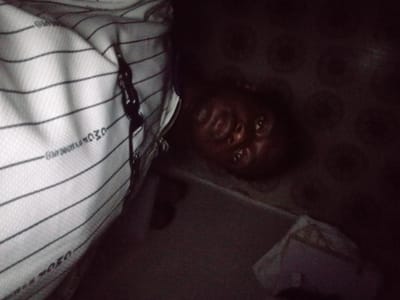 This dimly lit image, potentially taken indoors in Ogbogbo, Nigeria, features a person's face and a striped bag. The lighting is very poor, suggesting it was captured at night or in a room with minimal illumination.
The primary subject is a person with dark skin, whose face appears in the lower-right portion of the frame. Their head is tilted, and they are looking directly upwards towards the camera with open eyes. Due to the severe darkness, their expression is difficult to discern, but they appear awake.
To the left, a white fabric item with prominent horizontal black stripes dominates a large section of the image. This appears to be a bag, possibly a travel bag or backpack, as indicated by a visible black zipper running vertically along its right edge. Upon closer inspection, several words are faintly visible on the striped fabric. "OWO" can be seen near the top of the bag, and "OZOR" is visible upside down towards the bottom. Other smaller, less legible text may also be present, possibly related to a brand or item description.
The background is uniformly dark, appearing to be a floor or a solid surface, possibly with a subtle pattern in some areas. The camera perspective is unusual, suggesting a high angle looking down, or the subject might be lying down. There is no discernible activity or specific event taking place beyond the subject looking at the camera. The overall scene is casual and candid, characterized by its low light and intimate framing.