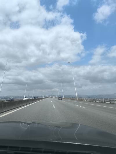 The image shows a view from a vehicle driving on a multi-lane bridge. The bridge is lined with tall, slender lampposts. On the left side of the bridge, there is a concrete barrier and several cars are visible in the opposing lanes. On the right side of the bridge, a metal railing separates the roadway from a body of water. In the distance, a city skyline with buildings and wind turbines can be seen across the water. The sky is partly cloudy with patches of blue sky visible. The overall scene suggests a typical daytime commute on a large bridge. This bridge is likely the Vasco da Gama Bridge, which connects Lisbon and the municipality of Samouco in Portugal.