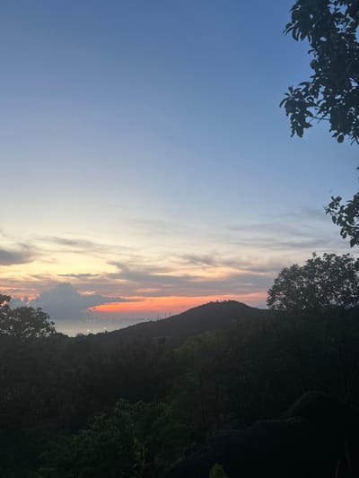 The image shows a view from Baan Hin Kong, Thailand, during a sunset or sunrise. The sky has colors ranging from blue to orange and pink, with light clouds. Dark tree foliage is visible in the foreground along the top and right side of the image. In the distance is a dark mountain ridge with what appear to be lights on the slope.