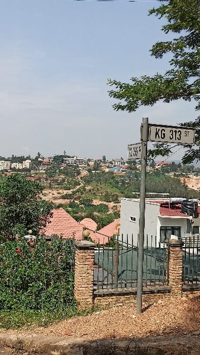 The image displays an elevated view of an urban landscape in Kigali City, Rwanda, under a clear or lightly hazy sky. In the foreground, a concrete curb borders a patch of dirt and gravel. A grey metal street signpost stands prominently, featuring two white rectangular signs: "KG 313 ST" pointing to the right, and "KG 355 ST" pointing to the left. To the left of the post, a dense green bush with a red flower is visible. Directly behind the post, a property is enclosed by a brick fence topped with dark green metal railings and decorative spherical light fixtures. A light-colored building with a reddish-brown tiled roof and two black water tanks on its roof is partially visible behind this fence. The mid-ground reveals a valley and opposing hills, densely covered with green vegetation and numerous residential buildings, primarily with red-tiled roofs. Patches of exposed reddish-brown earth are visible on the slopes. The distant background shows a more concentrated cluster of multi-story buildings atop the furthest hills. No people are depicted.
