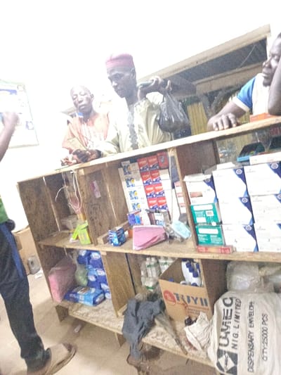 An indoor scene shows two men, one with a red hat, standing behind a wooden counter stocked with various items. The man in the red hat holds a dark plastic bag. Another man stands to the right of the frame. Below the counter, items like packaged sanitary pads, bottled liquids, and a large white bag labeled "UMEC" are visible. The overall lighting is dim, making the scene appear slightly cluttered. A partial figure is seen to the left.