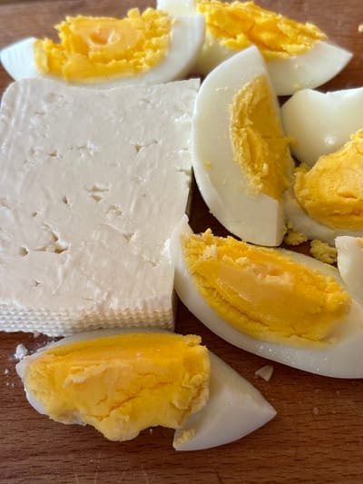 A close-up, overhead shot shows a block of white cheese next to several slices of hard-boiled eggs. The cheese is rectangular and has a textured surface with small holes. The eggs are cut in half lengthwise, revealing bright yellow yolks surrounded by white. The food is arranged on a wooden surface that appears to be a cutting board. The image is a still life, with no people or discernible action. The setting does not provide any information related to the city of Oreshak, Bulgaria.