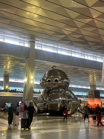 This image depicts a large, shiny, silver sculpture of a baby or child in what appears to be a modern, indoor public space, possibly an airport terminal, given the presence of people with luggage. The sculpture is the dominant subject, with its smooth, reflective surface mirroring the surroundings.
Several people are visible in the scene, most of whom are walking or standing with suitcases. Their attire suggests a casual, everyday setting. One person in the foreground wears a plaid shirt and dark pants, pulling a small suitcase. Further back, a person in a black shirt and jeans is also with luggage. Another individual is seen in a red top and pink pants.
The background features a geometrically patterned ceiling with triangular panels, illuminated by long strips of light. The floor is also reflective, showing the distorted reflections of the sculpture and people. Some greenery can be seen in planters on the right side of the image.
The lighting suggests it is either daytime with artificial lighting, or evening, given the bright interior lighting contrasting with any potential external light. There are no clear indications of weather conditions.
A sign with text is visible in the background, partially obscured but appearing to say "Selamat Jalan" which translates to "Goodbye" or "Have a safe journey" in Indonesian, reinforcing the idea of a travel hub. The overall atmosphere seems to be one of transit and everyday activity. The sculpture itself is a striking, almost whimsical element within this functional environment.