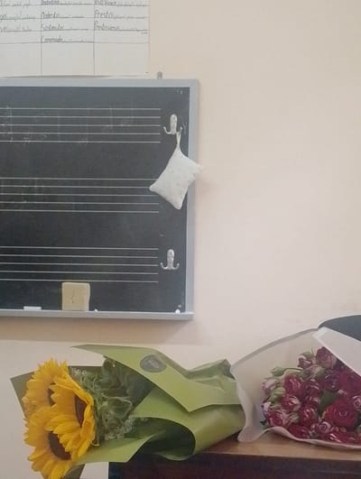 The image depicts a classroom setting with a chalkboard, musical notation, and two bouquets of flowers on a desk. The chalkboard has horizontal lines for musical staff, with some writing above it that appears to be musical tempo markings. There is a small white pillow attached to the top right of the chalkboard, and a piece of chalk or an eraser at the bottom left. A yellow chalk-like object is placed in the center of the bottom of the chalkboard. In the foreground, two bouquets of flowers are arranged on a wooden surface. The bouquet on the left consists of sunflowers, while the bouquet on the right is composed of small red roses. The flowers are partially wrapped in green and white paper. The background is a plain, light-colored wall. The overall scene suggests a school or educational environment, potentially a music classroom, decorated for a special occasion or as a gift. The city of Nor Hachin, Armenia, is not explicitly visible or indicated in the image itself; this information would need to be provided by an external source.