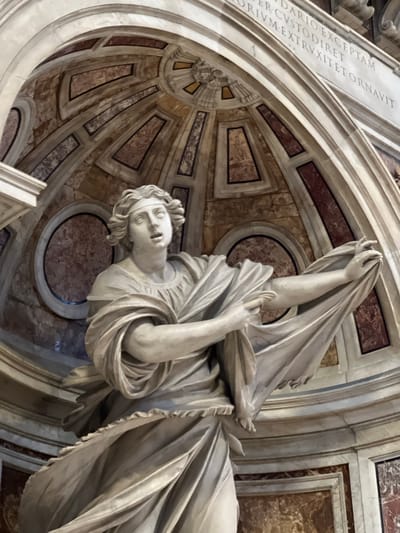 A white marble statue, depicting an allegorical female figure, is positioned prominently within an ornate architectural setting. The figure, identified as Justice from the Tomb of Pope Urban VIII, is shown in a dynamic pose, gazing upwards and to the left with an open mouth, conveying an intense expression. Her wavy hair is bound by a band. She wears classical drapery, with her left arm holding folds of fabric close to her body, while her right arm extends outwards, grasping a large section of billowing cloth.
The statue is set against a background of richly decorated architecture. Above the figure, a segment of a semicircular, coffered ceiling or dome is visible. This ceiling features alternating geometric panels (rectangular, trapezoidal) inlaid with reddish-brown and lighter veined marble, all outlined by white marble or stucco trim. A prominent white marble arch frames this upper section. Above the arch, a frieze bears a partial Latin inscription carved in capital letters, reading "...DARIO EXCEPTAM / ER CVSTODIRE ET / ORIVM EXTRVXIT ET ORNAVIT". Further into the background, several circular marble inlays are visible. The overall scene is characterized by the intricate detailing of marble in various colors, primarily white for the statue and structural elements, and shades of red, brown, and grey for the inlays. The location is the Tomb of Pope Urban VIII, situated in St. Peter's Basilica, Città del Vaticano, Vatican City.