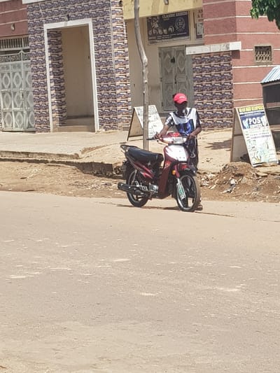 In Kufena, Nigeria, a person wearing a red cap and a striped t-shirt stands beside a red and black motorcycle on a dirt road. The motorcycle is parked on its kickstand. Behind the person and the motorcycle is a building with a patterned tile facade and an open doorway. To the right of the building, a sign with text and images is visible, and further to the right, another building with a red brick facade and a small, barred window is present. The ground is a mix of dirt and asphalt.