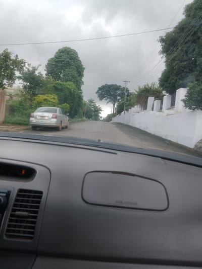 A silver sedan is parked on the right side of a paved road that curves upwards and to the right. The car faces away from the viewer and towards the right. A white wall with regularly spaced pillars runs along the right side of the road. Green foliage and trees are visible on both sides of the road, with a larger, mature tree on the left. The sky is overcast and grey. The scene is viewed from inside a vehicle, with the dashboard and part of the windshield visible in the foreground. The image appears to be taken in Iju, Nigeria.