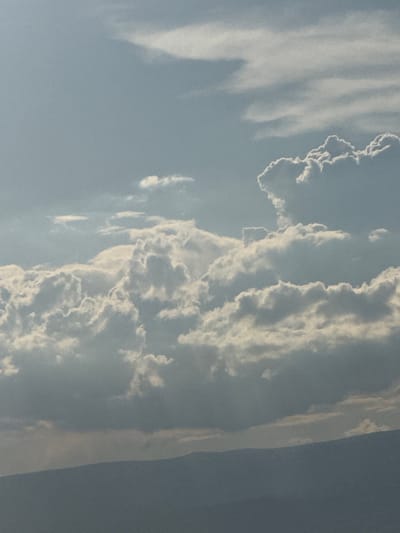 The image displays a wide shot of a hazy, blue sky filled with various cloud formations. In the foreground, the silhouette of a mountain range is visible, a dark blue-gray against the lighter sky. The clouds vary in density and appearance, with some appearing wispy and thin while others are thick and billowy, catching the light and casting shadows. A few scattered, smaller clouds are present in the upper portion of the frame. The overall impression is of a somewhat overcast or diffused lighting condition. No people, specific objects, or discernible actions are present in the image. The scene is devoid of any recognizable landmarks that would specifically indicate the city of Abovyan, Armenia.