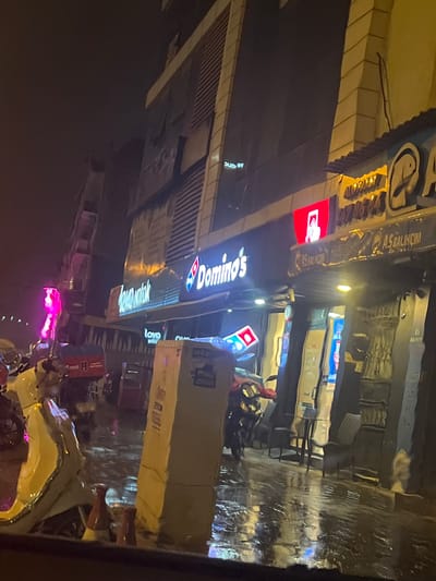 The image presents a nighttime urban street scene in Bayraklı, Turkey, characterized by wet, reflective pavement indicating recent or ongoing rainfall. A prominently lit Domino's Pizza storefront occupies the mid-ground, featuring its distinct blue and red logo. Directly above Domino's, a sign for "AS BALIKÇIM" (fish restaurant) is visible, with outdoor tables and chairs arranged on the wet sidewalk. To the left of Domino's, a sign for "love yatak" (a mattress store) is partially visible. A multi-story modern building with a glass and light-colored facade rises in the background. Several motorbikes and scooters are parked along the street or sidewalk; a cream-colored scooter is in the immediate foreground, and another with a red delivery box is further back near the Domino's entrance. A tall, light-colored rectangular utility box or pillar stands to the right of center. Artificial illumination from storefronts, streetlights, and a distinct pink neon light on the far left illuminates the scene, creating strong reflections on the wet surfaces. No individuals are clearly discernible.