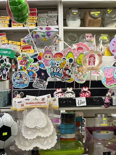 This image depicts a brightly lit interior of a commercial establishment, likely a party supply or general store, in the city of Partido de Quilmes, Argentina. The scene is dominated by a multi-tiered black display rack positioned centrally, showcasing numerous festive decorations.
In the upper section, a green and red striped foil balloon and a white polka-dotted foil balloon with "Feliz Cumple" (Happy Birthday) are visible, attached to sticks. Below these, a dense arrangement of stick-mounted cake toppers features various popular cartoon and media characters and themes. These include Super Mario, Bluey, Wednesday Addams, a pink rabbit, a "Feliz Día Papa Campeón del Mundo" (Happy Father's Day World Champion) topper with a soccer ball and Mickey Mouse, heart-shaped avocados, a "Mi Comunión" (My Communion) girl, Paw Patrol, and Peppa Pig.
On the lower tier of the black display, three small, heart-themed gift boxes are placed, two with red hearts on white and one with white hearts on brown. A small Minnie Mouse figurine stands to the right of these boxes. A white handwritten price tag displaying "1200" is also affixed to this tier.
Below the main display, transparent plastic packages containing white, heart-shaped lace doilies, labeled "BLONDA CORAZON," are visible. To the far left, a black and white soccer ball decoration is partially seen. Further to the right, a stack of colorful spools of ribbon (dark brown, blue, light blue, orange) is arranged.
The background features white shelving units. The top shelf contains several clear glass jars with metal lids, holding various bulk items such as white powder (possibly sugar or flour), a light brown powder (likely cinnamon), and other unidentified granular substances. The middle and lower shelves are cluttered with numerous cardboard boxes and containers, some with handwritten labels. Visible labels include names of Argentine soccer clubs such as "RIVER," "BOCA," "CELESTE," "RACING," and "SAN LORENZO," further indicating the geographical context of the store.