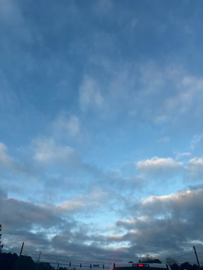 The image displays an outdoor scene in Lawrenceville, United States, focusing on the sky and elements of a street environment. The upper two-thirds of the frame consists of a light blue sky with scattered white and grey cloud formations. Across the mid-ground, multiple dark utility poles and overhead wires are visible, with dark tree lines extending beneath them. In the immediate foreground, the top-rear section of a light-colored pickup truck is partially visible, featuring a central red brake light illuminated on its tailgate. Above the truck, several dark traffic light fixtures are suspended from overhead wires. No individuals are present in the scene, and no discernible actions or interactions are occurring.