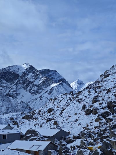 The image depicts a winter scene in what appears to be a mountainous region. Several buildings, possibly dwellings or lodging, are visible at the base of the frame, covered in snow. The landscape surrounding the buildings is rocky and also covered in snow. In the background, there are snow-covered mountains under a cloudy sky.
While the image context suggests a location in Khumjung, Nepal, as specified in the prompt, the picture itself does not provide any visible elements that specifically confirm the exact geographic coordinates or landmark to definitively corroborate that location.